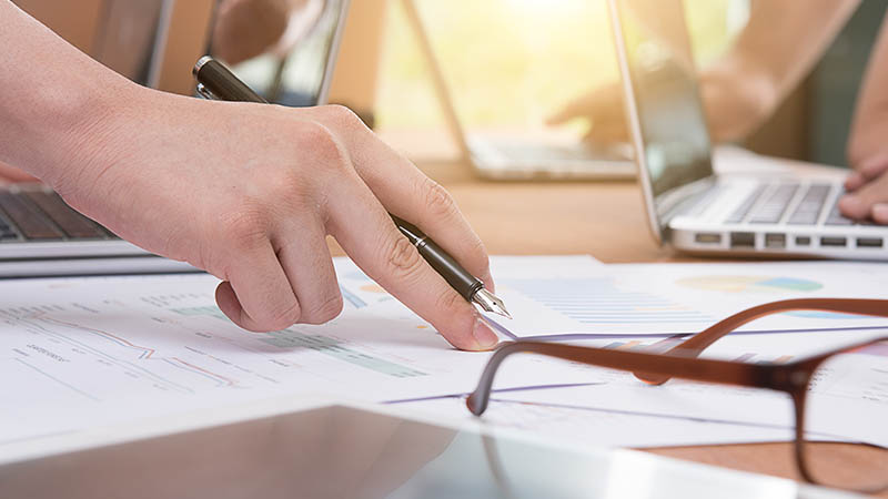 Papeles, lentes y computadoras en una mesa. Una mano en el centro, por escribir un papel
